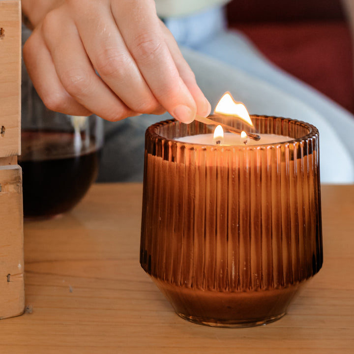 Hand lighting a candle in a ribbed brown glass holder on a wooden table.
