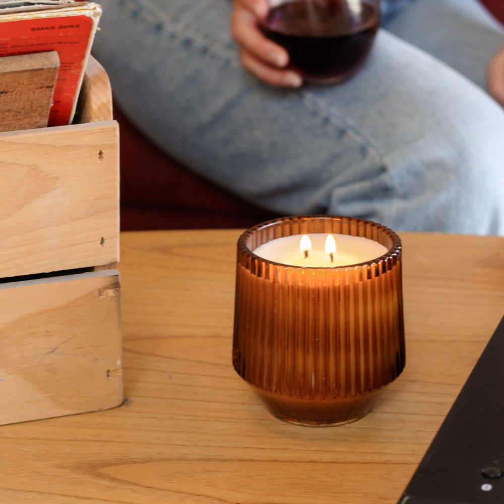 Candle in a textured brown holder on a wooden table with a person holding a glass of red wine in the background.