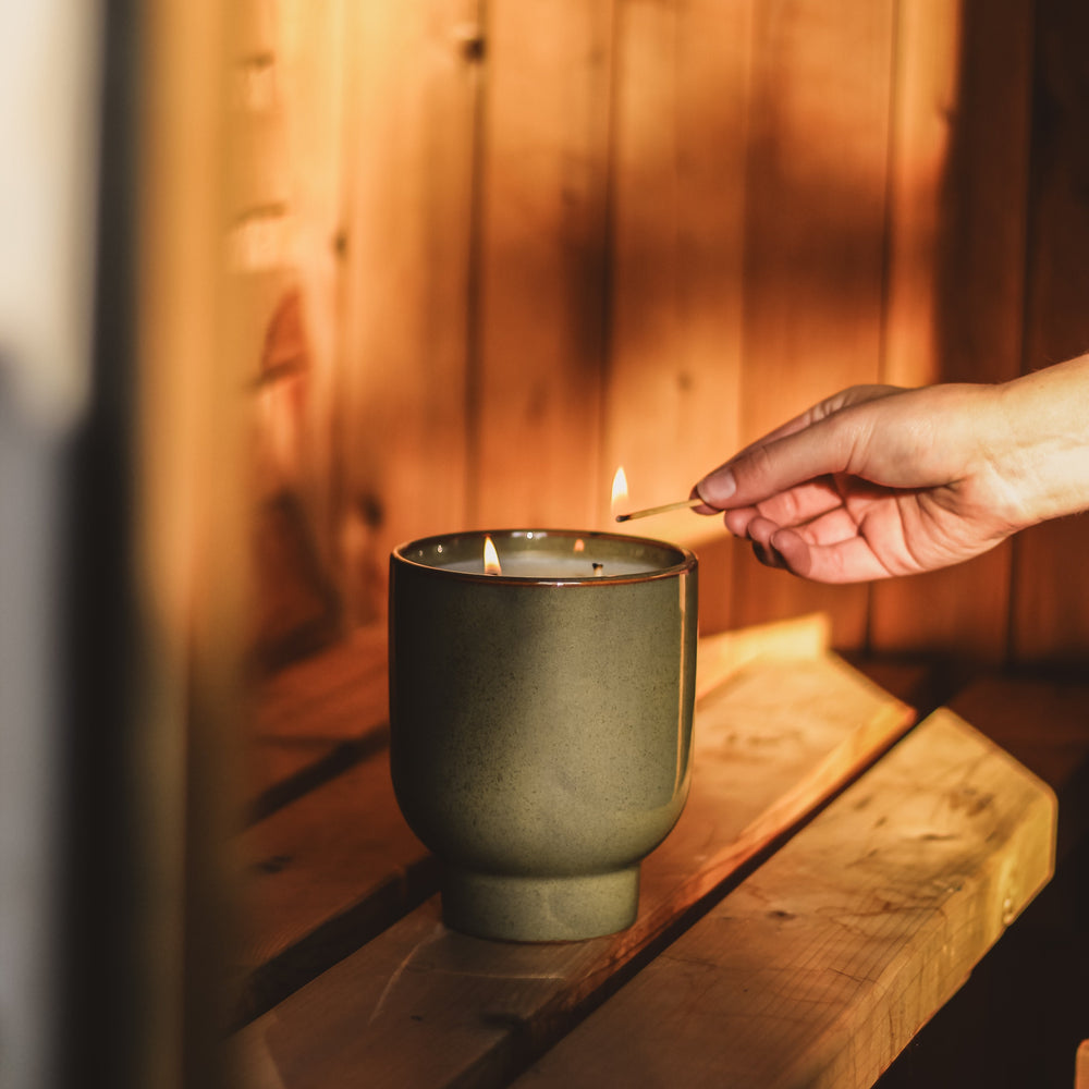 Hand lighting a candle in a green container on a wooden surface with a warm, wooden background.