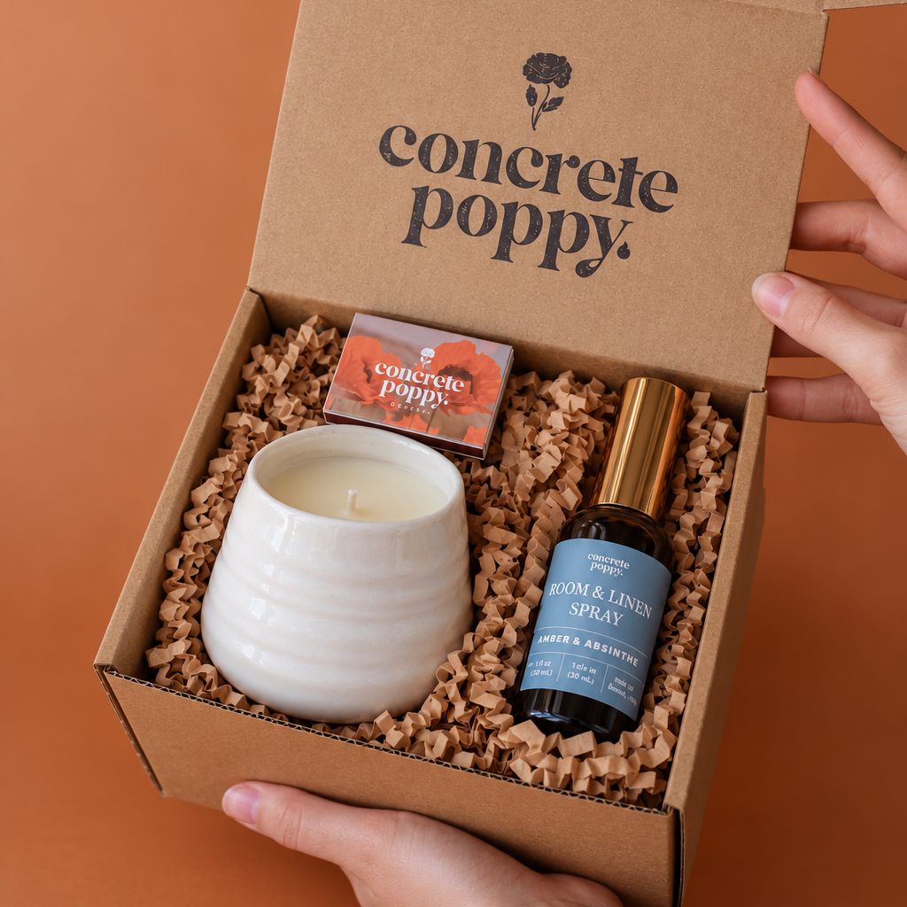 A gift box from Concrete Poppy held open by hands against a solid terracotta background. The kraft cardboard box is filled with brown packing material and contains a white ceramic candle, a small orange box, and a dark amber bottle with a blue label and gold cap.