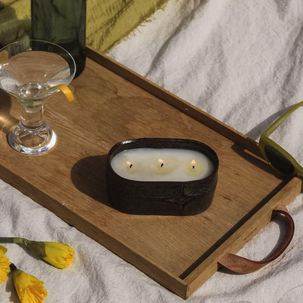 Candle on a wooden tray with a bottle, glass, and flowers on a white blanket.
