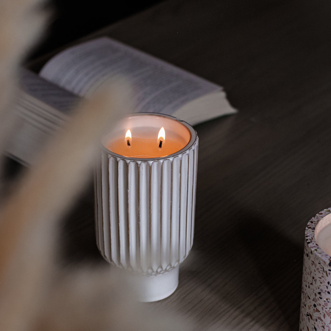 Ceramic Candle in a textured white holder on a wooden surface with an open book in the background