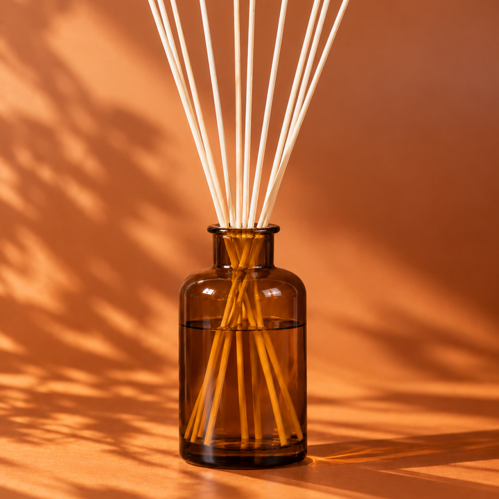 Amber glass reed diffuser bottle with natural wooden reeds, set against a warm orange background with soft leaf shadows.