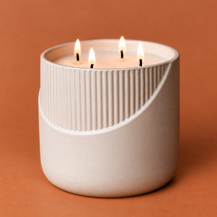 A modern three-wick candle in a ribbed ceramic container with a curved cutout, lit and set against a solid terracotta background.