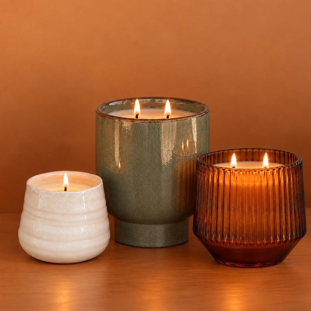 Three lit candles in decorative glass holders on a wooden surface against a warm brown background. The candles include a small white ribbed ceramic holder, a medium-sized textured sage green holder, and a larger amber brown ribbed glass holder.