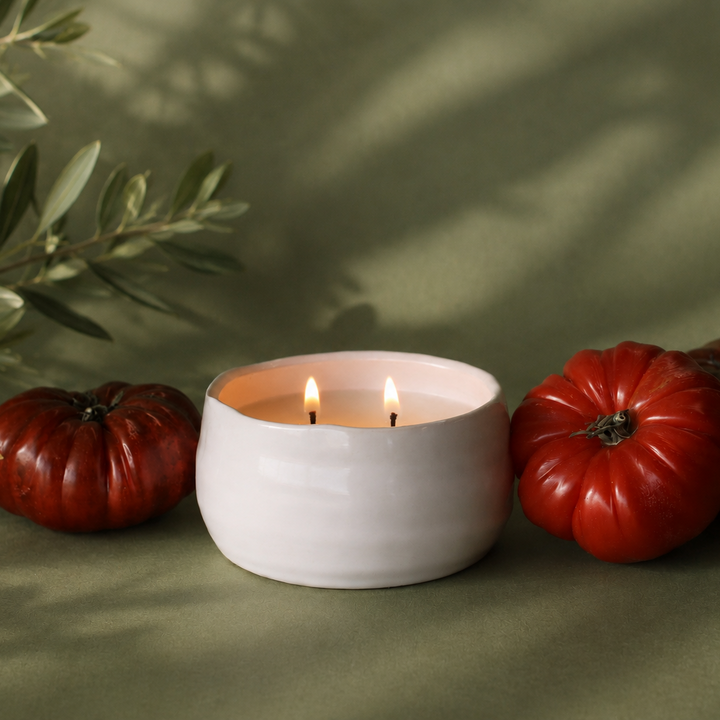 White ceramic candle holder with a lit candle, surrounded by red tomatoes and green leaves on a green background.