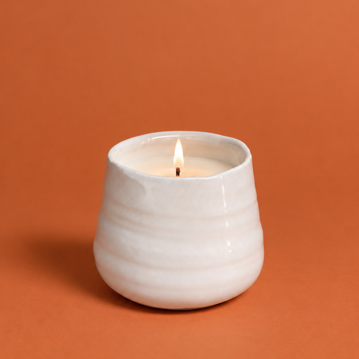 A lit white ceramic candle holder with a single wick, set against a solid terracotta orange background.